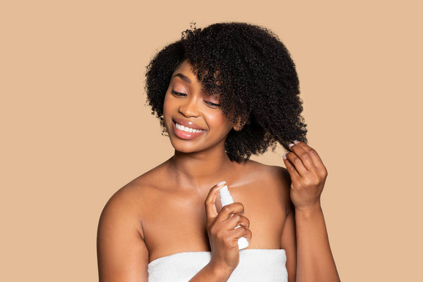 Vidám fiatal fekete nő törölközőben alkalmazza tápláló hajápoló termék a göndör haj, mosolyogva kellemesen krémes bézs háttér - Fotó, kép