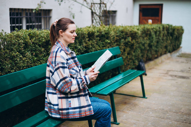 Piękna stylowa kobieta w modnym nosić spędzać czas na ulicy przy użyciu papierowej mapy. Szczęśliwa dziewczyna siedząca na ławce. Koncepcja miejskiego stylu życia, baner turystyczny - Zdjęcie, obraz