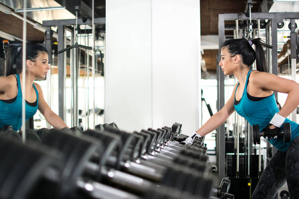 Mujer joven eligiendo pesas en el gimnasio. Se queda cerca de un espejo y ve su reflejo - Foto, imagen