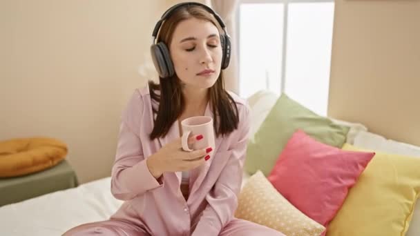Een beschouwende jonge vrouw in pyjama met hoofdtelefoon en een koffiemok zit op een bed versierd met kleurrijke kussens in een gezellige slaapkamer. - Video