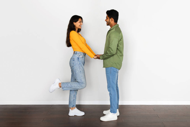 Sorrindo jovem casal hindu brincando de mãos dadas, mulher levantando a perna em pose de paquera, de pé contra a parede branca, mostrando momento romântico - Foto, Imagem
