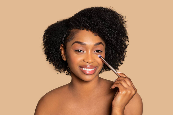 Mujer negra brillante con rizos naturales aplica cuidadosamente corrector debajo de su ojo con cepillo de maquillaje, disfrutar de la rutina de belleza contra el fondo beige - Foto, imagen
