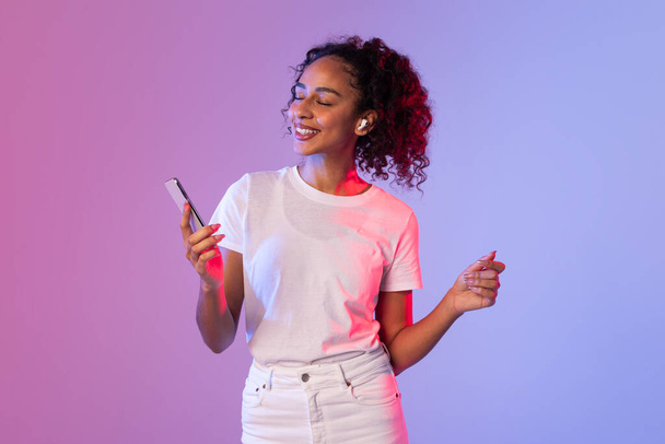 In un vivace sfondo al neon, una donna con i capelli ricci tiene uno smartphone mentre fa un gesto alla mano - Foto, immagini