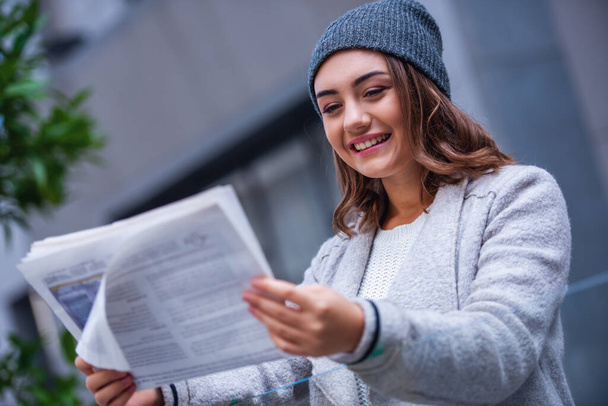 Красивая девушка в пальто и кепке читает газету и улыбается во время отдыха на открытом воздухе
 - Фото, изображение