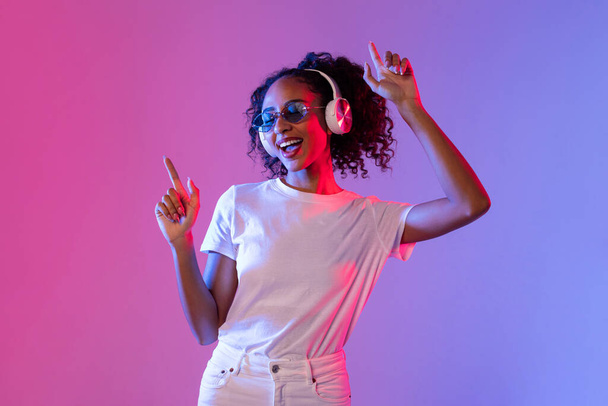 Vibrant young black woman with curly hair enjoying music in wireless headphones and dancing against pink and blue gradient neon backdrop - Photo, Image