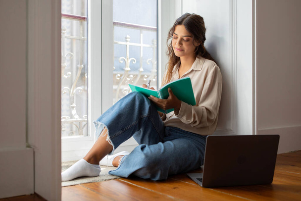 Focada jovem mulher escrevendo em notebook enquanto sentado no chão de madeira perto de laptop por janela brilhante em casa, bela fêmea milenar estudando em ambiente doméstico confortável, espaço de cópia - Foto, Imagem