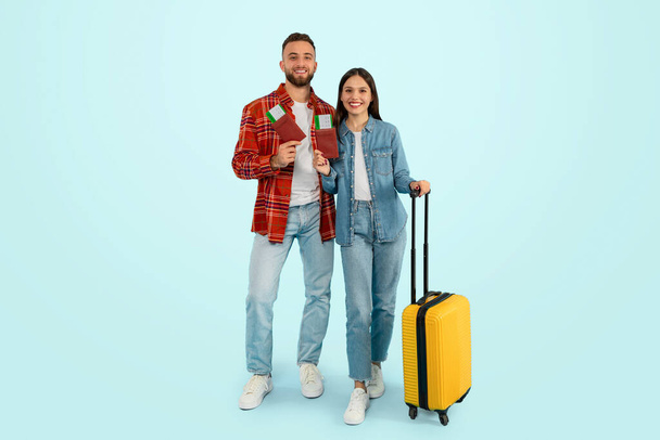 Wesoły kaukascy podróżnicy para ekscytująco czeka na wymarzone wakacje, stojąc z biletami i walizką, uśmiechając się, jak planują romantyczny wypad, pozowanie na niebieskim tle. Pełna długość - Zdjęcie, obraz