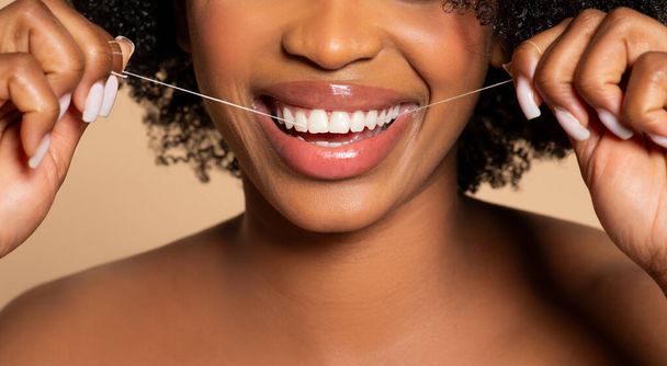 Primo piano del sorriso radioso come donna nera con bei capelli ricci filo interdentale sapientemente i denti, promuovendo l'igiene orale su sfondo neutro - Foto, immagini