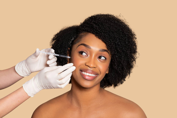 Νεαρή μαύρη γυναίκα με λαμπερό βλέμμα που λαμβάνει ένεση υαλουρονικού οξέος, προσβλέπει σε ενισχυμένη λάμψη του δέρματος σε απαλό μπεζ φόντο - Φωτογραφία, εικόνα