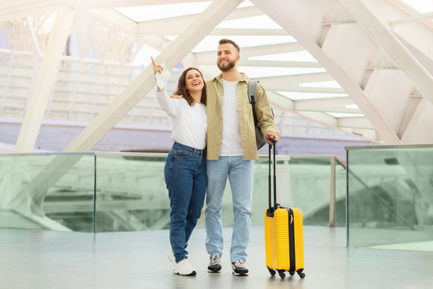 Glückliches junges Paar genießt seine Zeit am Flughafen beim Warten auf den Flug, fröhliche Millennials mit gelbem Gepäck in der Terminalhalle, Frau zeigt weg und lächelt - Foto, Bild