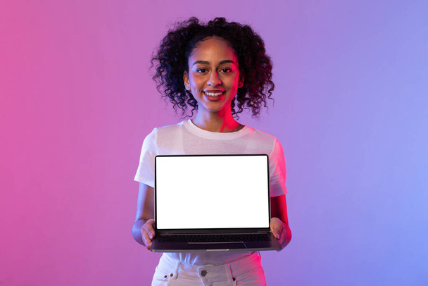 Hymyilevä musta kihara tukka nainen tilalla avoin kannettava tietokone valkokankaalla valmis brändäys, seisoo vastaan violetti ja vaaleanpunainen kaltevuus neon tausta - Valokuva, kuva