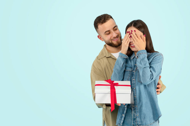 Celebrating Valentines Day. Man playfully offering present box while his wife covers eyes, standing against blue studio background, sharing cherished moment of celebration together. Copy space - Photo, Image