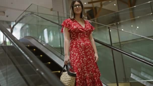 Alegre hermosa mujer hispana con gafas posa en escalera mecánica del centro comercial, irradiando un aura positiva mientras compras en el centro urbano - Metraje, vídeo