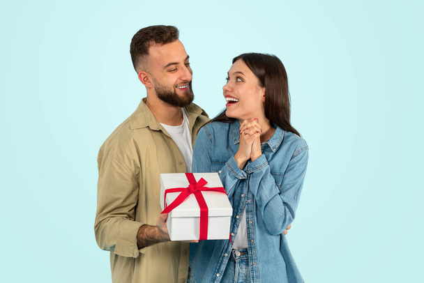 陽気な白人男性は,青いスタジオの背景に彼の妻を祝福する包まれたプレゼントボックスを与えます. 一緒にロマンチックな休日を祝う若いカップル. 記念日のコンセプト,誕生日サプライズ - 写真・画像