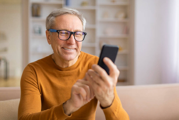 Gözlüklü, gülümseyen yaşlı bir adam akıllı telefonuna odaklanmış ev ortamında ilgi ve rahatlık taşıyor. - Fotoğraf, Görsel