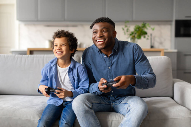 Un moment joyeux quand un jeune garçon et son père s'engagent dans une compétition ludique de jeux vidéo sur le canapé - Photo, image