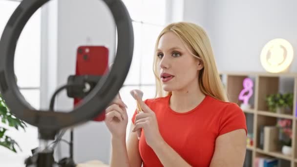 Szőke nő alkalmazó smink, miközben rögzíti a tutorial egy fényes stúdióban egy gyűrű fény és okostelefon. - Felvétel, videó