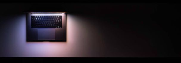 Laptop z świecącym ekranem na stole w ciemności, widok z góry. Sieć, szablon banera mediów społecznościowych. Przestrzeń kopiowania. - Zdjęcie, obraz