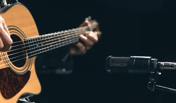 Mužský hudebník hrající na akustickou kytaru za mikrofonem v nahrávacím studiu. Koncept nahrávání hudby, zkoušení nebo živého vystoupení. - Fotografie, Obrázek