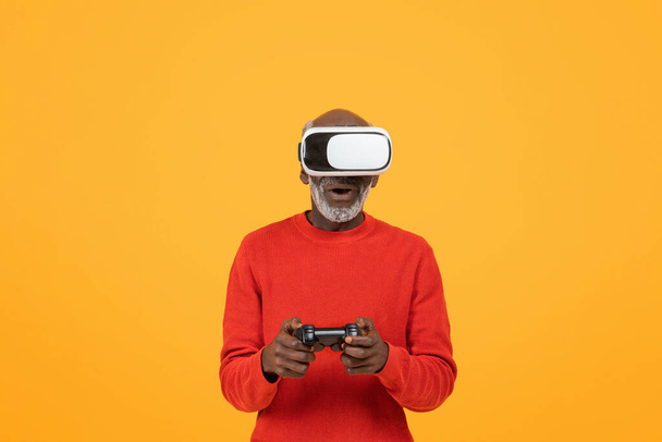 Incrível feliz homem negro sênior vestindo um fone de ouvido realidade virtual e segurando um controlador de jogo, totalmente imerso em uma experiência VR interativa em um fundo amarelo, estúdio - Foto, Imagem