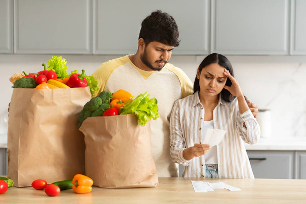 Frustré malheureux couple indien millénaire avec des sacs en papier plein d'aliments biologiques vérifier les factures après l'épicerie, conjoints choqués par les prix élevés, intérieur de la cuisine confortable - Photo, image