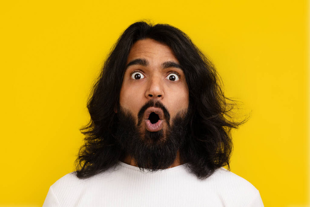 Portrait of shocked bearded long-haired millennial indian guy with eyes and mouth wide open posing on yellow studio background, expressing emotions, closeup - Photo, Image