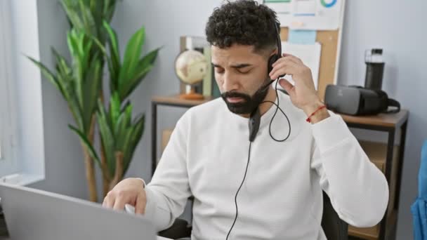 A bearded young hispanic man in headphones gesturing while working in a modern office interior. - Footage, Video
