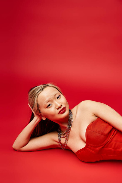 Uma mulher asiática atraente em um vestido vermelho graciosamente descansa no chão. - Foto, Imagem