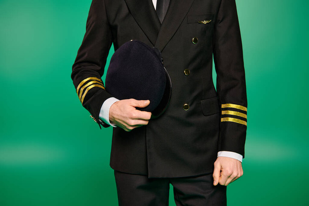 Guapo piloto masculino con uniforme negro sosteniendo un sombrero sobre un fondo verde. - Foto, Imagen