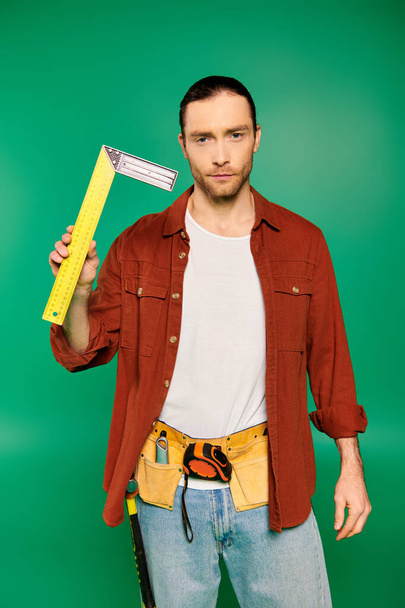 Bonito trabalhador masculino em uniforme segurando uma grande fita métrica amarela contra um pano de fundo verde. - Foto, Imagem