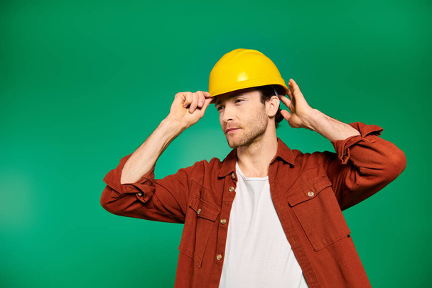Красивий працівник чоловічої статі в уніформі вражає позу на зеленому тлі з жовтим твердим капелюхом. - Фото, зображення