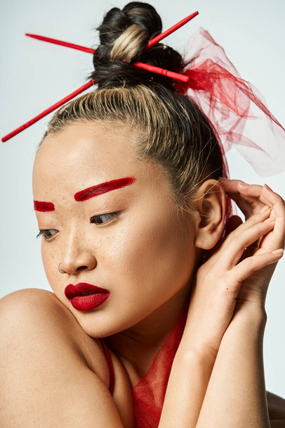 Femme asiatique vibrante avec du maquillage rouge et des bâtons dans ses cheveux, frappant une pose puissante. - Photo, image