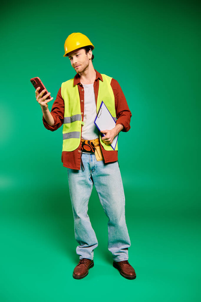 Ένας όμορφος εργάτης με κίτρινο γιλέκο ασφαλείας κρατά με αυτοπεποίθηση ένα κινητό τηλέφωνο. - Φωτογραφία, εικόνα