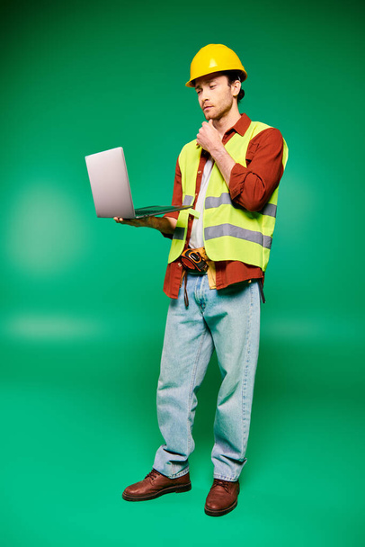 Handsome worker in hard hat holding laptop on green backdrop. - Photo, Image