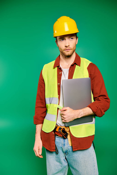 Kvalifikovaný pracovník v klobouku sebevědomě drží notebook v zeleném pozadí. - Fotografie, Obrázek