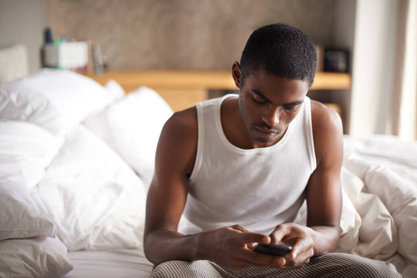 Schwarzer Mann, Schreibmaschine und Bett mit Telefon für Kommunikation, soziale Medien oder Netzwerkarbeit zu Hause. Afrikanische männliche Person am Morgen auf dem Handy für Online-Chat, SMS oder App im Schlafzimmer zu Hause. - Foto, Bild