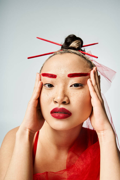 Mujer asiática en traje vívido, con maquillaje rojo, sostiene su cabeza en una pose dramática. - Foto, Imagen