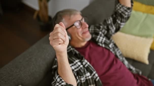 Egy vidám, ősz hajú férfi szemüveget visel, nevet, miközben a kanapén fekszik egy otthonos belső térben.. - Felvétel, videó