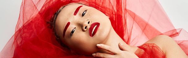 Uma mulher asiática cativante com maquiagem vermelha vibrante e um véu vermelho fluindo posa graciosamente. - Foto, Imagem