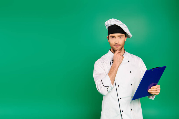 Guapo chef masculino en uniforme blanco sosteniendo un portapapeles sobre fondo verde. - Foto, imagen