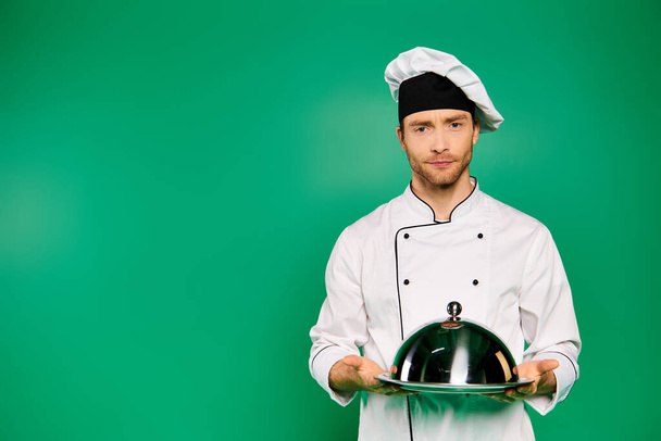 Ein gutaussehender männlicher Koch in weißer Uniform hält stolz einen Teller vor grünem Hintergrund. - Foto, Bild