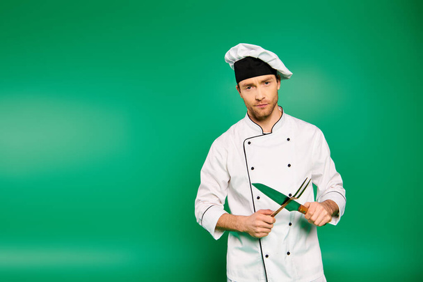 Fešák samec kuchař v bílé uniformě obratně drží příbory na zeleném pozadí. - Fotografie, Obrázek