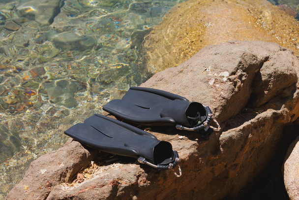 Twee zwarte zwemvinnen voor snorkelen of duiken rusten op een rotsachtig oppervlak naast helder water. Ze zijn naast elkaar geplaatst. Zonlicht verlicht de rotsen en helderheid van het water. Een flippers. - Foto, afbeelding