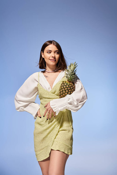 Beautiful young woman in a flowing dress gracefully holds a ripe pineapple in a vibrant studio setting. - Photo, Image