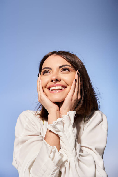 Une jeune femme aux cheveux bruns sourit joyeusement, les mains reposant doucement sur son visage dans un décor de studio. - Photo, image