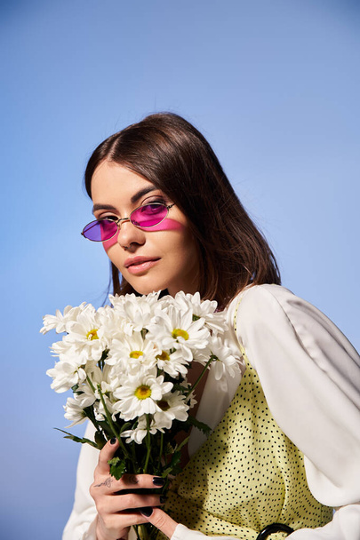 Молода жінка з брюнеткою, одягнена в сонцезахисні окуляри, тримає букет ромашок в безтурботній студії. - Фото, зображення