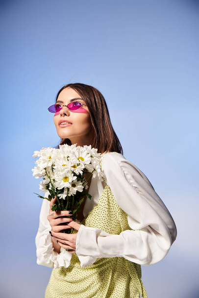 Eine junge Frau mit brünetten Haaren trägt eine Sonnenbrille und hält einen Strauß Gänseblümchen in einem ruhigen Studio-Ambiente. - Foto, Bild