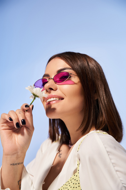 Eine elegante junge Frau mit brünetten Haaren trägt eine Sonnenbrille und hält eine lebendige Blume in der Hand. - Foto, Bild