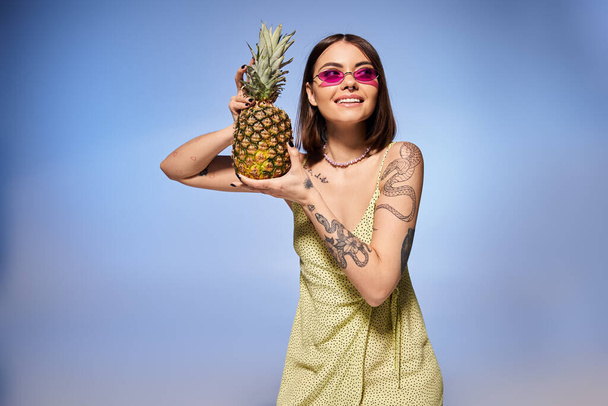 A young woman with brunette hair elegantly poses in a vibrant yellow dress, holding a fresh pineapple. - Fotó, kép