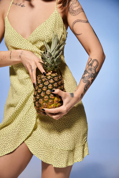 Egy barna hajú fiatal nő áll elegánsan sárga ruhában, friss ananásszal a kezében.. - Fotó, kép